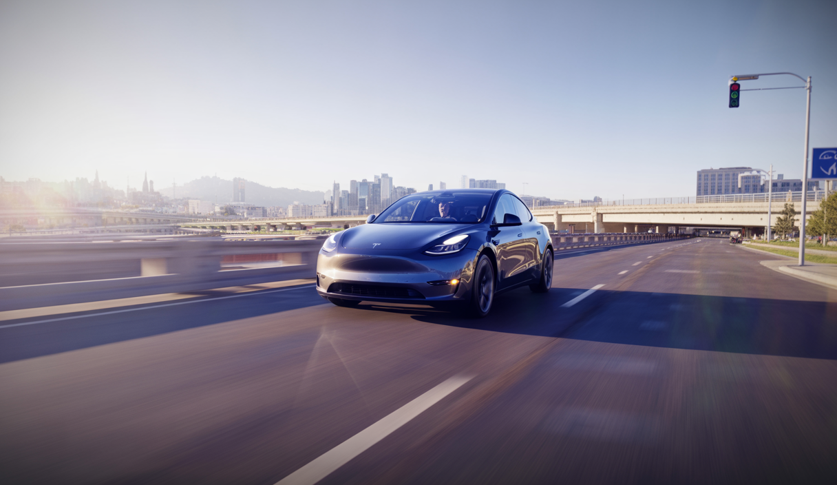 Tesla Model Y 2025 en conduite entièrement autonome durant sa première livraison officielle de l'usine au domicile du client, montrant la voiture circulant seule sur une route urbaine moderne sous un ciel clair